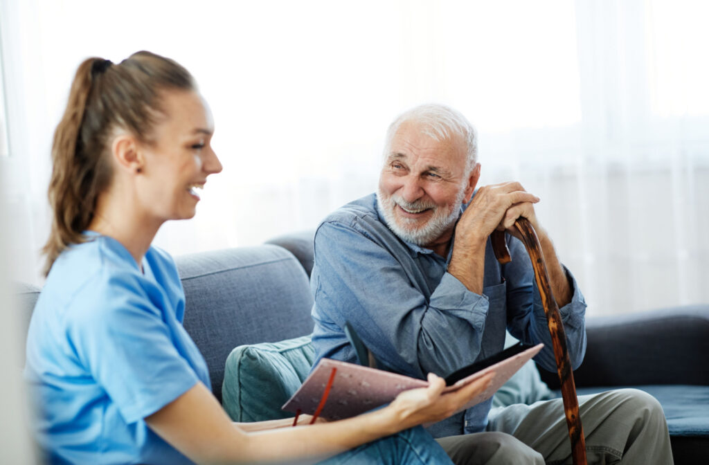 An older adult man sitting with a cane conversing with a nurse holding a book.