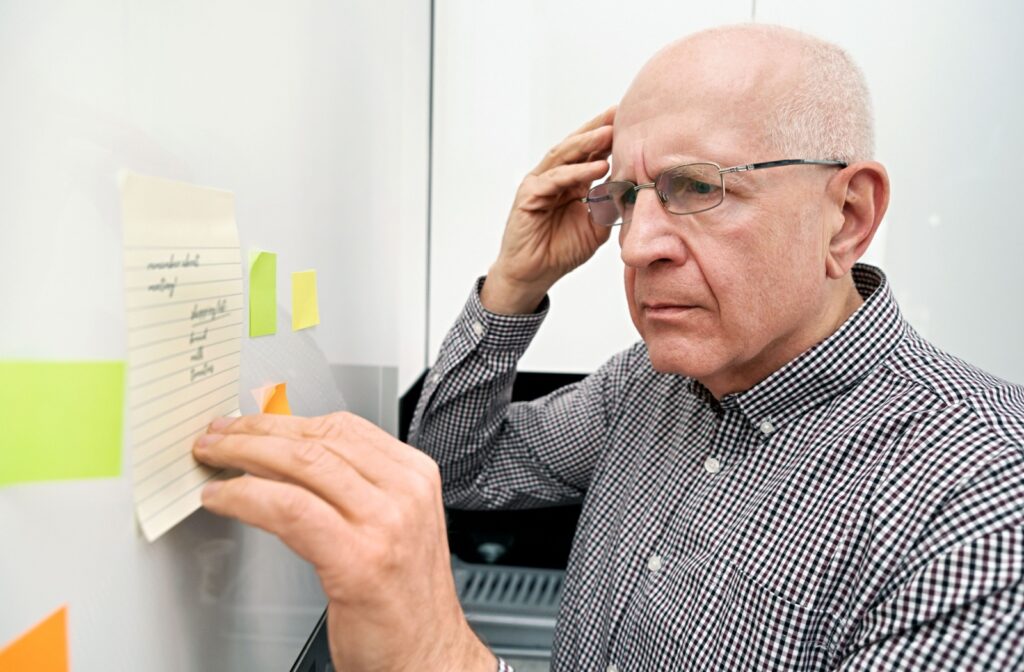 An elderly man looks at a note on the wall and touches his head in confusion.